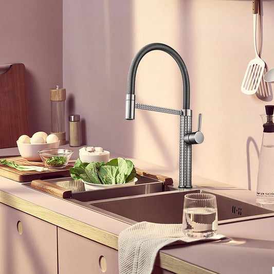 A sunlit kitchen features the Tiqui Windsor Solid Brass Pull‑Down Kitchen Faucet with a high-arc swivel spout, leafy greens in the sink, a glass of water on the counter, and assorted utensils and vegetables in the background.