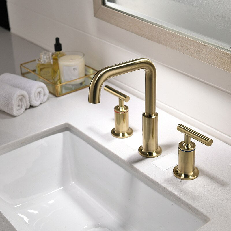The Tiqui Arched Twin-Handle Deck-Mount Bathroom Faucet sits on a white countertop, paired with matching handles. A rolled white towel and toiletries in a tray are beside the sink, with a mirror above.