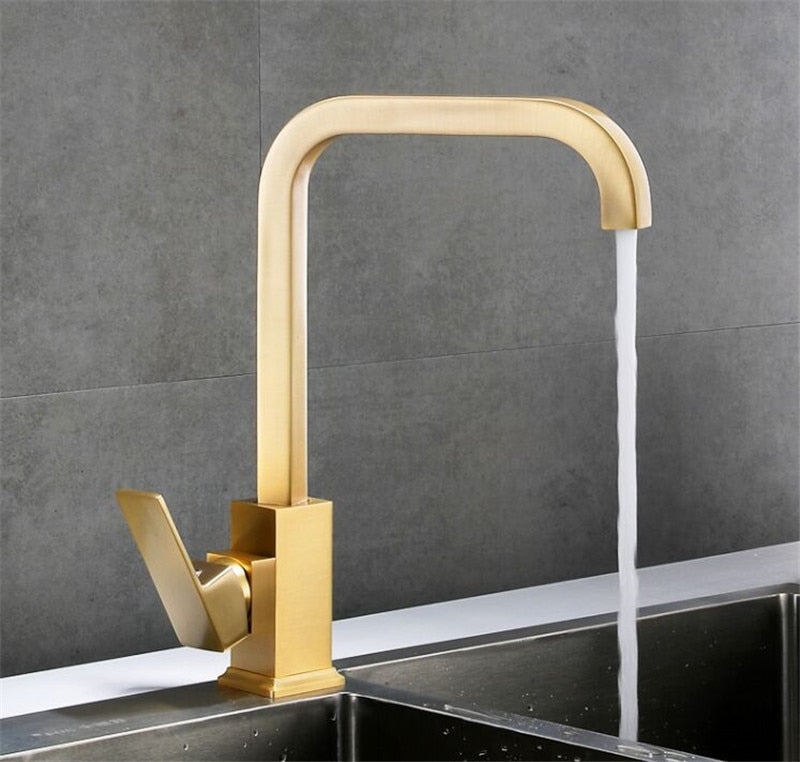 The Tiqui™ Solid Brass Single Handle Kitchen Faucet by Tiqui, featuring a sleek, modern golden finish and a square design, is shown mounted on a sink against a dark, tiled wall backdrop. Water is flowing from the faucet into the sink, and it has a lever handle on its side.