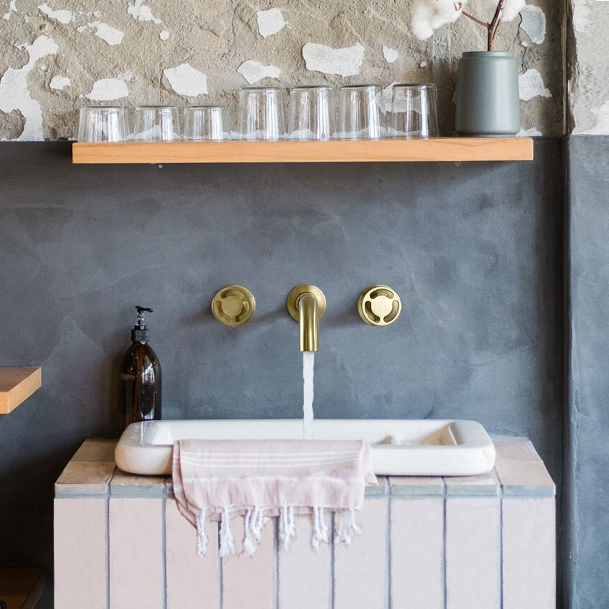 A Solid Brass Dual Handle Wall Mounted Faucet from Morsale.com, featuring two round handles and a curved spout, is installed above a bathtub. Reminiscent of a luxurious shower head design, this brass faucet releases water into the tub while a towel is partially visible at the edge. The background showcases a textured, light gray wall.