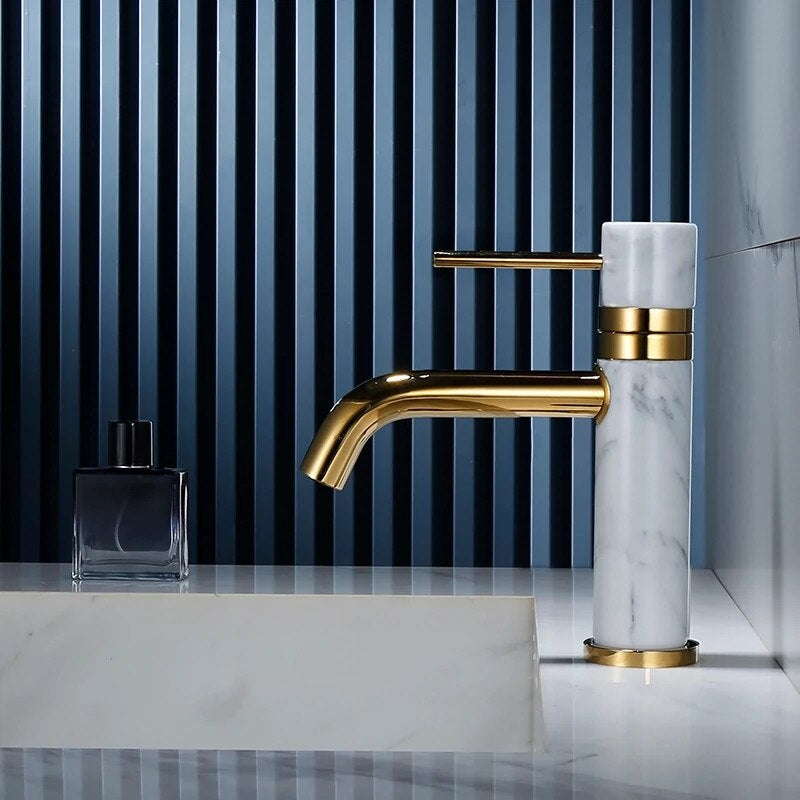 The Solid Brass & Marble Bathroom Faucet by allfixture enhances a modern bathroom sink with its sleek, gold finish and marble handle, placed beside a small square bottle of dark-colored liquid. The backdrop of vertically aligned blue and black slats perfectly complements the minimalist and sophisticated design of the bathroom faucet.