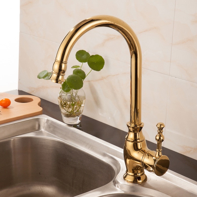 A Tiqui Polished Gold Kitchen Faucet with a curved spout is installed above a stainless steel sink. In the background, a small glass vase with green leaves and a wooden cutting board with a cherry tomato enhance the scene.