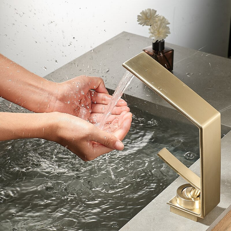 A modern Brushed Gold Solid Brass Bathroom Sink Faucet by Morsale.com is mounted on a gray countertop, with water flowing gently from its rectangular spout into the sink below. Next to the faucet, there is a small vase with white flowers and a black square container.