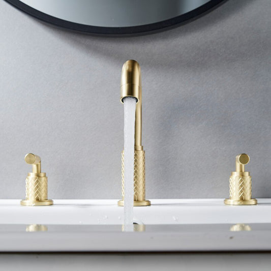 The Windsor Widespread Bathroom Sink Faucet by Mike Jake features dual handles and a solid brass build, with water flowing into a white basin set against a light gray wall and part of a round mirror visible above.