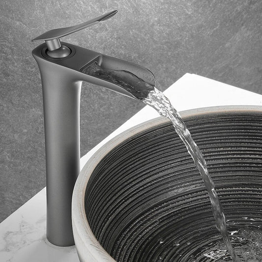 The Tiqui Valmont Single Handle Bathroom Sink Faucet, in matte gray gunmetal with a flat spout, pours into a round dark-striped ceramic sink set on a white marble countertop against a gray textured wall.
