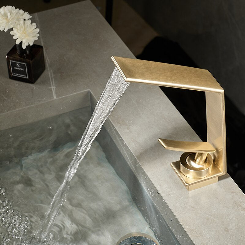 A modern Brushed Gold Solid Brass Bathroom Sink Faucet by Morsale.com is mounted on a gray countertop, with water flowing gently from its rectangular spout into the sink below. Next to the faucet, there is a small vase with white flowers and a black square container.
