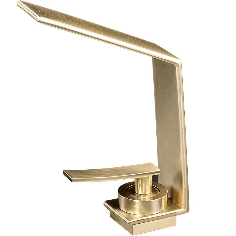 A modern Brushed Gold Solid Brass Bathroom Sink Faucet by Morsale.com is mounted on a gray countertop, with water flowing gently from its rectangular spout into the sink below. Next to the faucet, there is a small vase with white flowers and a black square container.