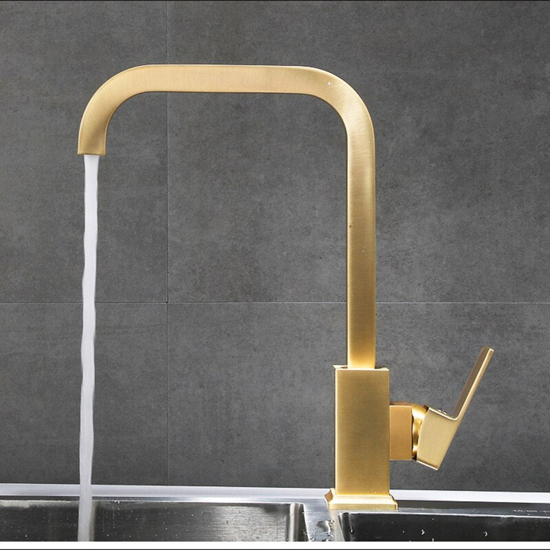 The Tiqui Solid Brass Single-Handle Kitchen Faucet features a modern gold finish, high arc spout, and square design, releasing a steady stream into a stainless steel sink set before a gray tiled wall.