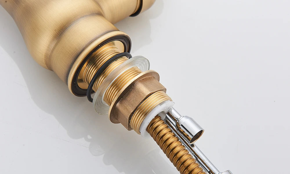 A close-up of the Rozin Antique Brass Pull-Down Spout Kitchen Faucet with a high arc and a pull-down sprayer. A hand is shown holding the sprayer, with water flowing into a stainless steel sink. A white curtain and a white bowl with purple flowers are visible in the background, enhancing the kitchen's charm.