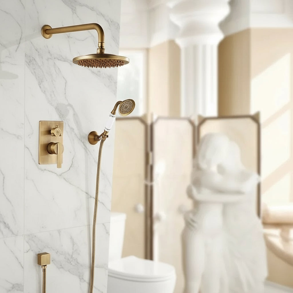 The Rozin Heritage Complete Shower System, featuring gold solid brass fixtures, is mounted on a marble-tiled wall. In the background are a toilet and a blurred white statue of embracing figures.