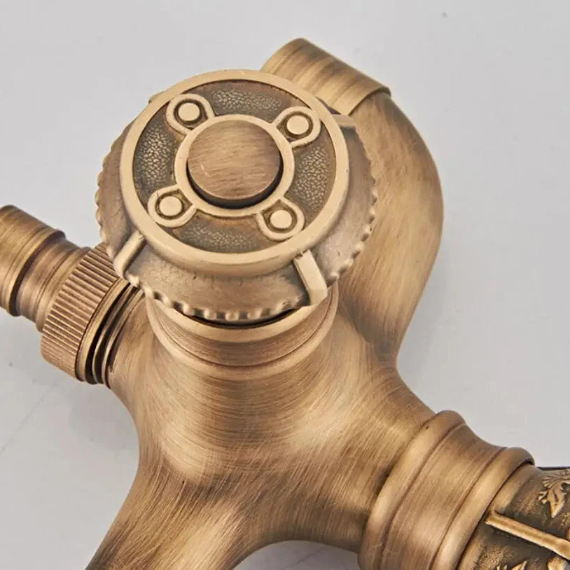 Close-up of the Allfixture Antique Brass Bidet Faucet Wall Mounted Carved Bidet Faucet, showcasing an intricate, circular handle. The wall-mounted faucet features detailed engravings and a rustic finish that impart an antique appearance. The valve has three connection points and textured ridges for grip and control, pairing beautifully with an ornate shower head.
