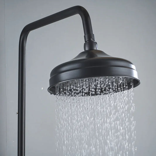The Rozin Heritage Complete Shower System features a round black solid brass showerhead mounted on a pipe, releasing a steady stream of water droplets against a plain gray background.