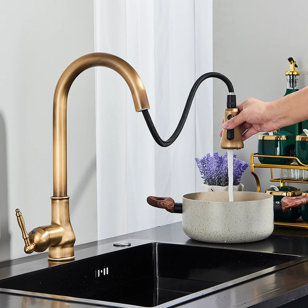 A close-up of the Rozin Antique Brass Pull-Down Spout Kitchen Faucet with a high arc and a pull-down sprayer. A hand is shown holding the sprayer, with water flowing into a stainless steel sink. A white curtain and a white bowl with purple flowers are visible in the background, enhancing the kitchen's charm.