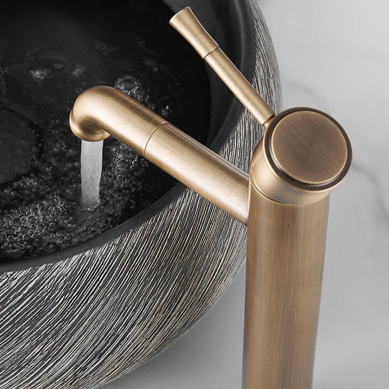 An Antique Bronze Single Handle Bathroom Sink Faucet from Tiqui stands tall beside a round, textured stone sink with a grey and white swirl pattern. Complementing the design is a sleek shower faucet in the background against the textured, stone-like grey wall, creating a stylish and minimalist bathroom aesthetic.