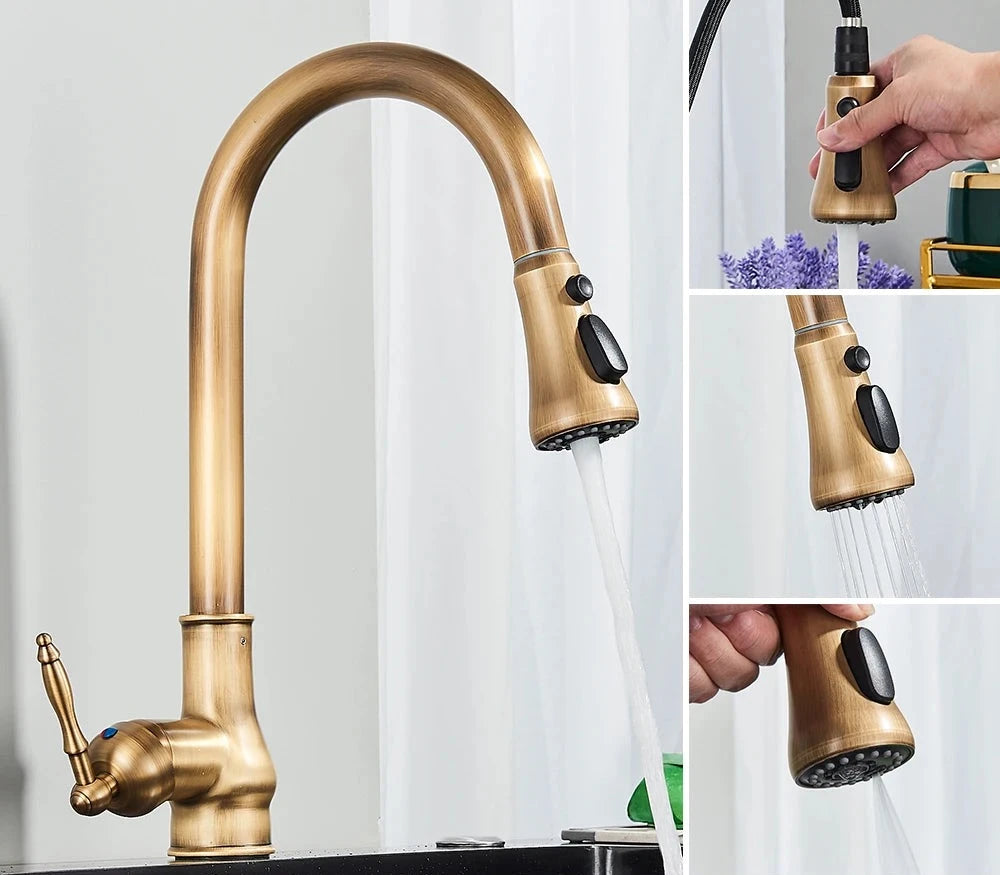 A close-up of the Rozin Antique Brass Pull-Down Spout Kitchen Faucet with a high arc and a pull-down sprayer. A hand is shown holding the sprayer, with water flowing into a stainless steel sink. A white curtain and a white bowl with purple flowers are visible in the background, enhancing the kitchen's charm.