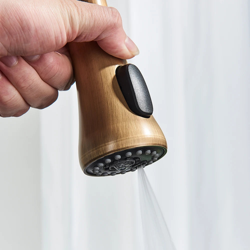 A close-up of the Rozin Antique Brass Pull-Down Spout Kitchen Faucet with a high arc and a pull-down sprayer. A hand is shown holding the sprayer, with water flowing into a stainless steel sink. A white curtain and a white bowl with purple flowers are visible in the background, enhancing the kitchen's charm.
