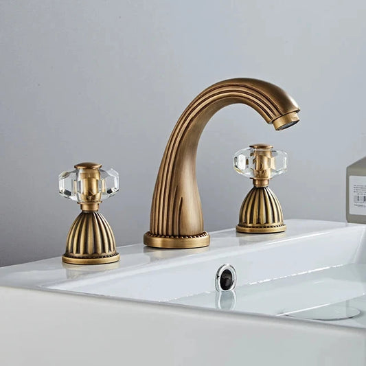 The Allfixture Heritage Widespread Basin Faucet features a gold-tone, 3-hole design with two crystal-style clear faceted handles, shown installed on a modern white sink against a clean light gray background.