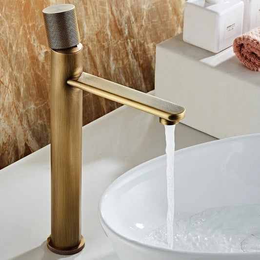 The Tiqui Maison Antique Brass Bathroom Faucet features a modern, textured handle and gold-toned finish, dispensing water into a white round sink set on a bathroom counter with a brown marble wall and neatly arranged toiletries.