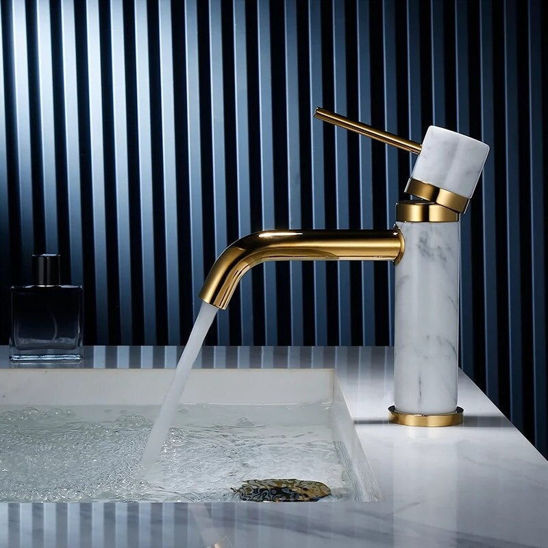The Solid Brass & Marble Bathroom Faucet by allfixture enhances a modern bathroom sink with its sleek, gold finish and marble handle, placed beside a small square bottle of dark-colored liquid. The backdrop of vertically aligned blue and black slats perfectly complements the minimalist and sophisticated design of the bathroom faucet.