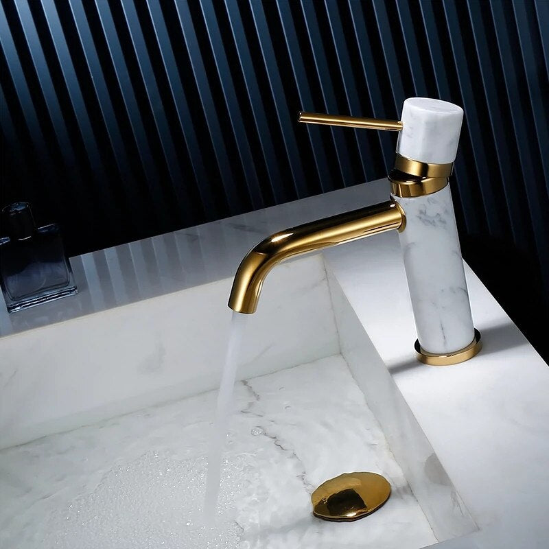 The Solid Brass & Marble Bathroom Faucet by allfixture enhances a modern bathroom sink with its sleek, gold finish and marble handle, placed beside a small square bottle of dark-colored liquid. The backdrop of vertically aligned blue and black slats perfectly complements the minimalist and sophisticated design of the bathroom faucet.