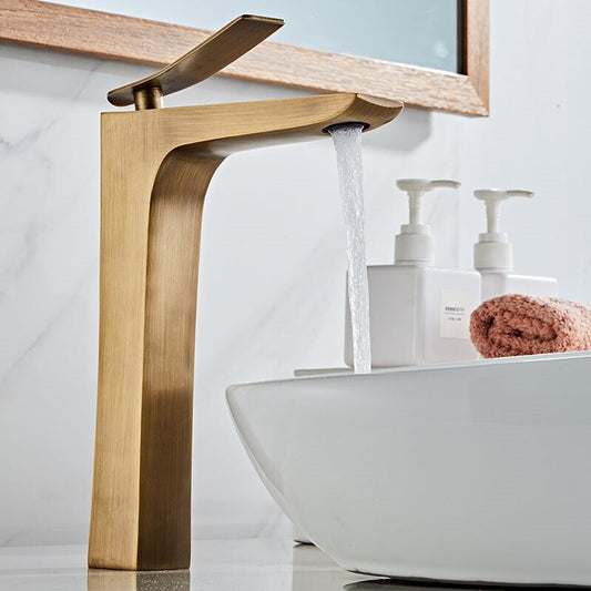 The Tiqui Windsor Deck Mounted Bathroom Sink Faucet, featuring a flat lever spout in modern gold, pours into a white vessel sink; a pink towel and two soap dispensers rest on the surrounding white marble countertop.