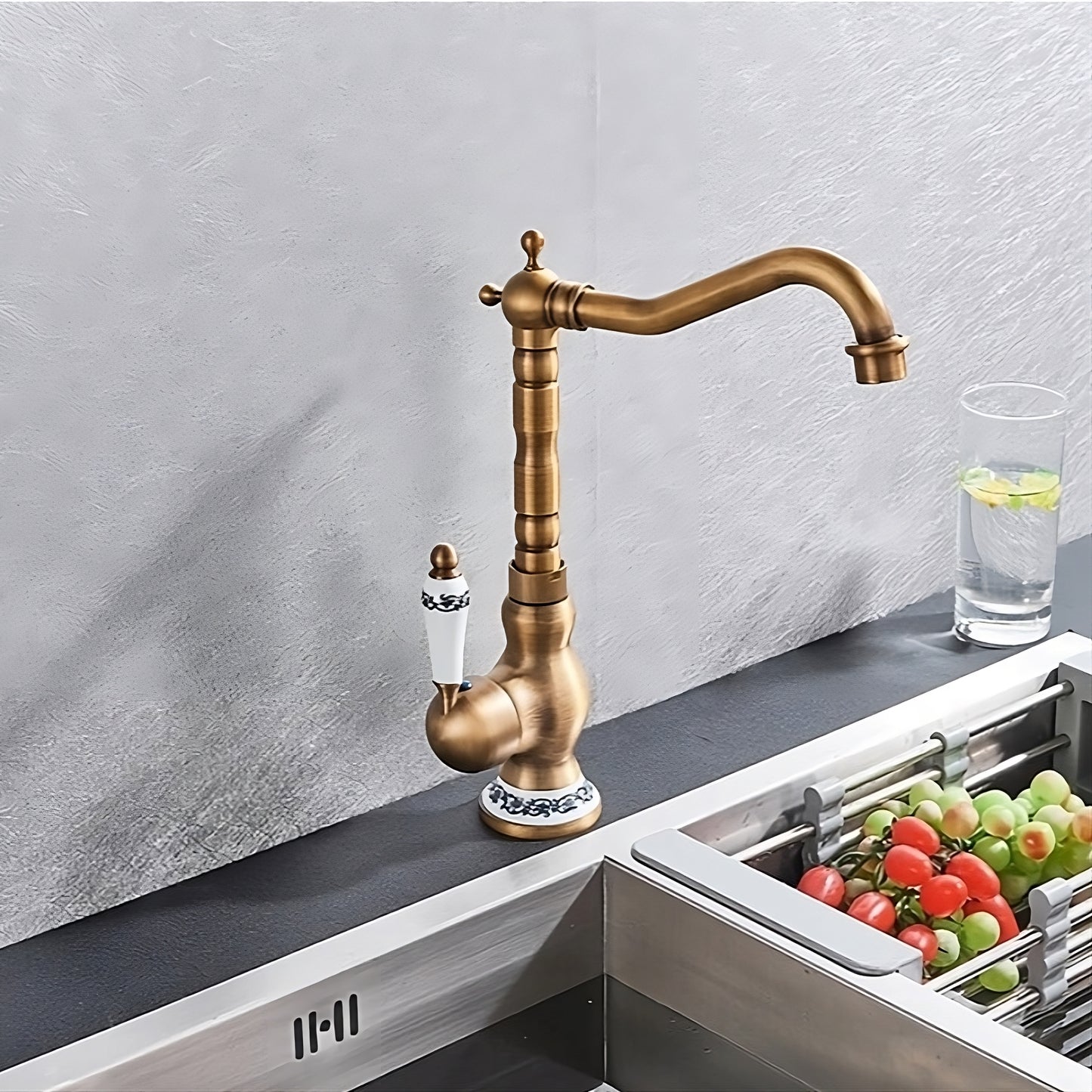 The Ellen Antique Brass Single-Handle Kitchen Faucet, featuring a high-arc spout and decorative handle, is installed on a countertop beside a sink with a glass of water and fresh tomatoes and cucumbers.