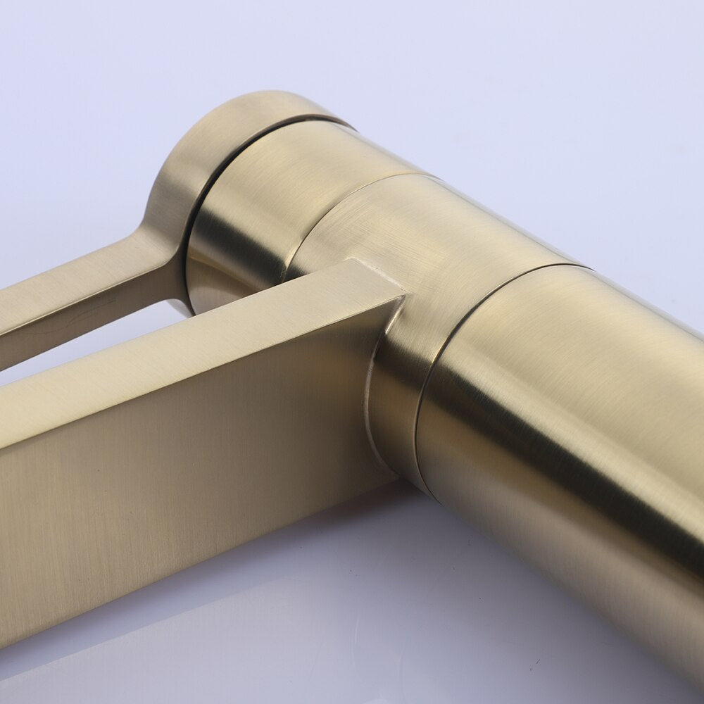 A modern ROZIN Brushed Gold Single Handle Faucet by allfixture is mounted on a white bathroom sink, with water flowing from it. A folded beige towel is placed on the left corner of the sink. The background features white tiled walls, adding a touch of elegance akin to a sophisticated kitchen faucet.