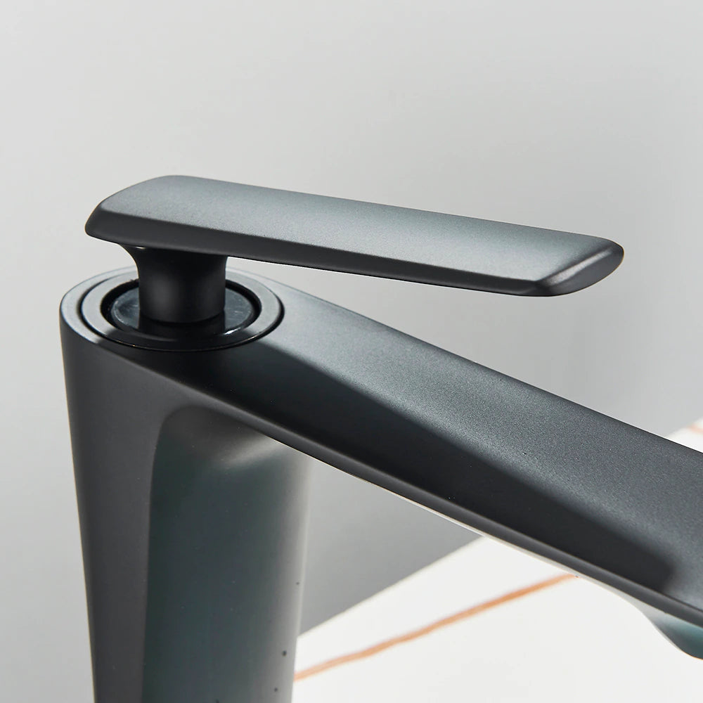 A Wasser™ Solid Brass Bathroom Sink Faucet by Rozin, featuring a sleek, angular design in modern black, dispenses water into a round, white vessel sink. In the background, a partial reflection is seen in a circular mirror, while a small plant adds a touch of greenery to the scene.