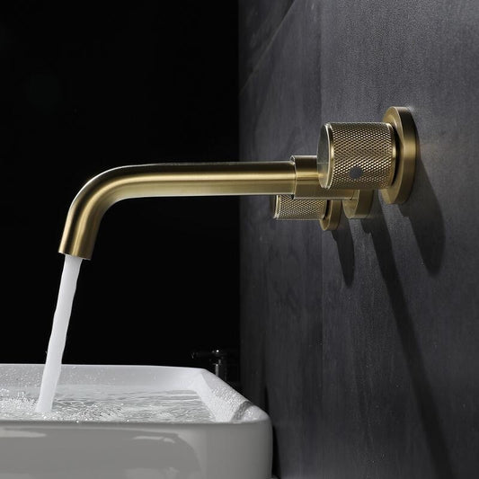 The Tiqui Solid Brass Wall Mounted Bathroom Faucet in brushed gold, which features two round handles and a stylish spout, pours water into a white rectangular sink filled with water against a dark gray tiled wall.