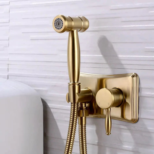 The AllFixture Wasser™ Solid Brass Hot & Cold Water Bidet Faucet, featuring a gold finish and matching wall mount with control knobs, is installed on a white tiled bathroom wall with its flexible hose coiled neatly below the handheld spray.