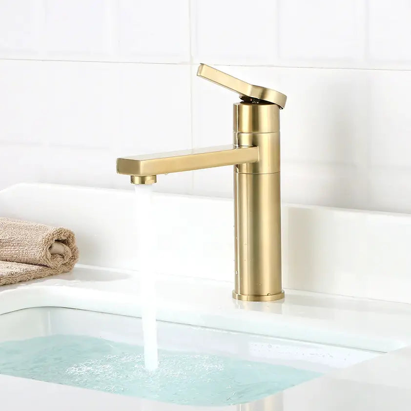A modern ROZIN Brushed Gold Single Handle Faucet by allfixture is mounted on a white bathroom sink, with water flowing from it. A folded beige towel is placed on the left corner of the sink. The background features white tiled walls, adding a touch of elegance akin to a sophisticated kitchen faucet.