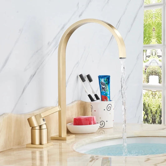 A brushed gold solid brass deck mounted bathroom sink faucet by allfixture showcases a modern high-arc spout and a single lever handle. Installed on a white countertop with a blurred plant and mirror in the background, this faucet creates a cohesive contemporary look, seamlessly complementing any kitchen sink faucet or shower head in matching finishes.