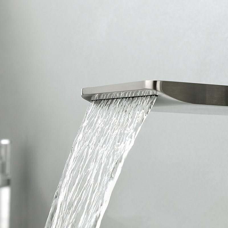 The Sekova Wall Mounted Bathtub Faucet With Handheld Shower Sprayer is depicted. It features a modern design with a sleek, rectangular spout that allows water to flow smoothly. Adjacent to the spout, on the same panel, is a handheld showerhead equipped with a hose. Both the fixture and the surrounding wall boast a brushed stainless steel finish.