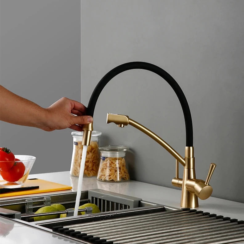 A person is using the Wasser™ Solid Brass Kitchen Faucet With Filtered Water Tap by allfixture to wash green apples in a sink. The countertop features a cutting board with a tomato, and jars of pasta are visible in the background. The overall kitchen decor is minimalist and contemporary, highlighting the modern design of the sleek gold and black kitchen faucet.