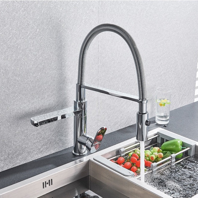 A sleek AllFixture Deck Mounted Swivel Spout Pull Down Kitchen Faucet with a flexible, high-arc, stainless steel spring spout is shown installed over a stainless steel sink. Water flows from the faucet into the sink, washing various fresh vegetables. This design brings the modern elegance often reserved for bathroom faucets into your kitchen.