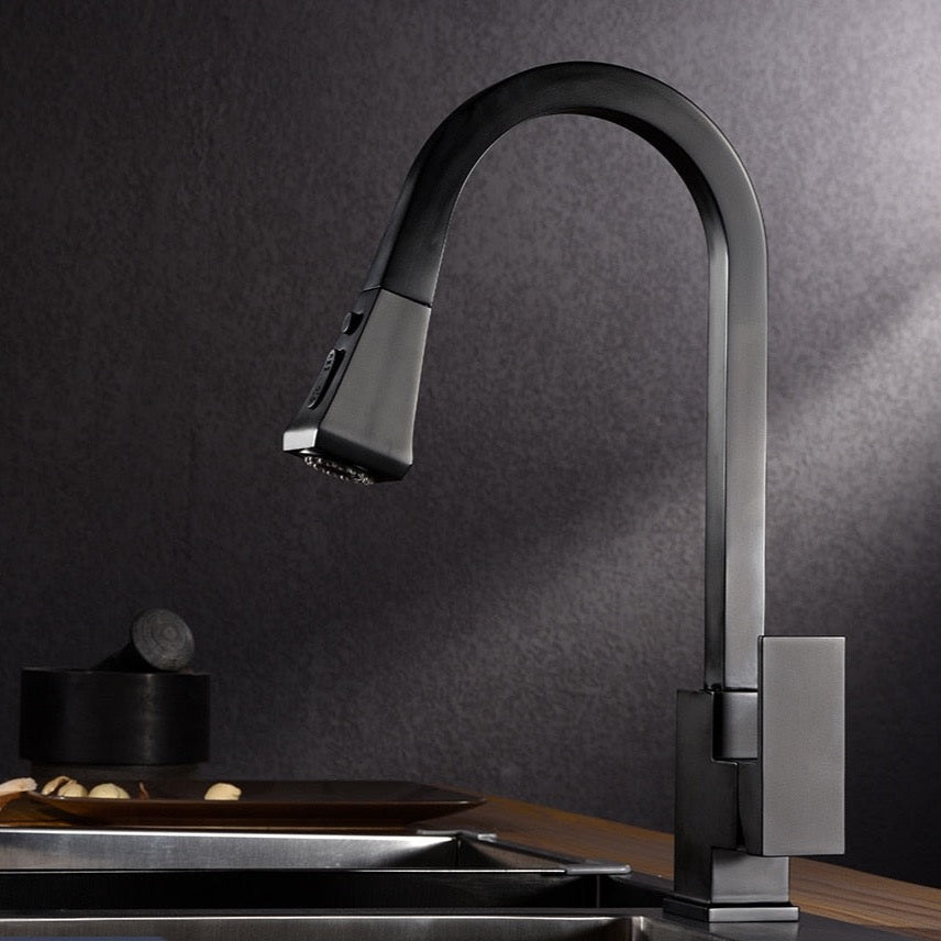 A modern kitchen features the Wanfan Solid Brass Pull Out Kitchen Faucet by allfixture, characterized by its sleek, high-arc chrome design. The contrasting aesthetic is created by a black backsplash and a white countertop. Two wine glasses and a white bowl filled with oranges are placed on the counter, illuminated by natural light from a nearby window.
