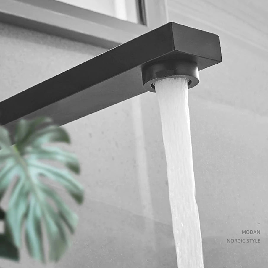 An AllFixture Wall Mount Swivel Spout Wash Basin Bathroom Faucet, featuring a modern, chrome-plated design with minimalist elements, pours a steady stream of water against a light gray tiled wall. The faucet has a sleek, rectangular form that enhances the contemporary aesthetic of the bathroom setting.