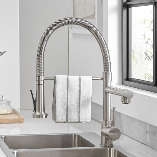 The allfixture Wanfan™ Dual Spout Kitchen Faucet, with its sleek, modern stainless steel design and handy pull-down sprayer, is dispensing water into a glass pitcher. The faucet is perfectly positioned in front of a large window that offers a beautiful view of lush greenery.
