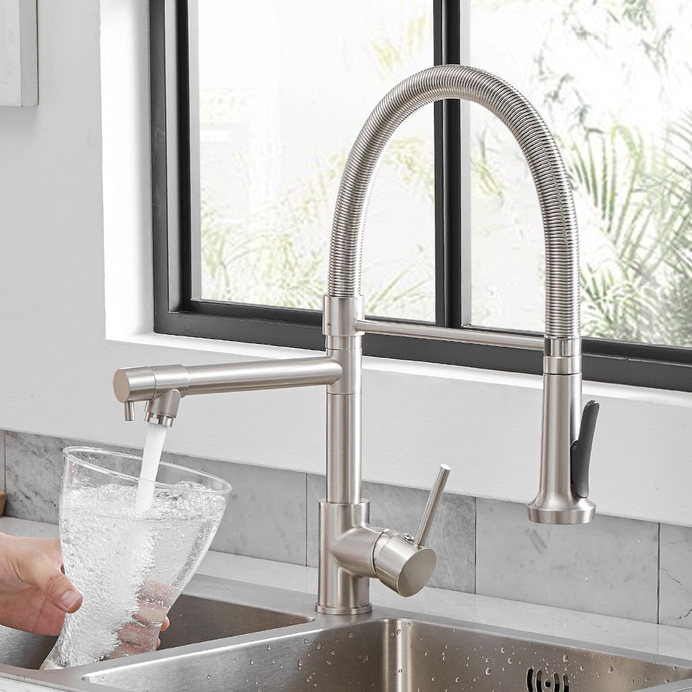 The allfixture Wanfan™ Dual Spout Kitchen Faucet, with its sleek, modern stainless steel design and handy pull-down sprayer, is dispensing water into a glass pitcher. The faucet is perfectly positioned in front of a large window that offers a beautiful view of lush greenery.