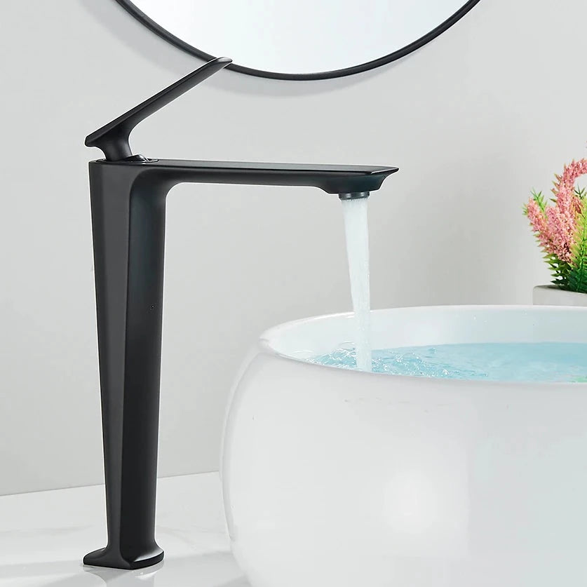 A Wasser™ Solid Brass Bathroom Sink Faucet by Rozin, featuring a sleek, angular design in modern black, dispenses water into a round, white vessel sink. In the background, a partial reflection is seen in a circular mirror, while a small plant adds a touch of greenery to the scene.
