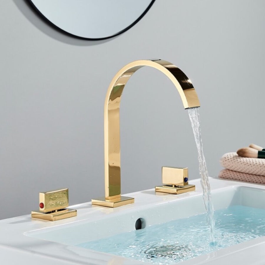 A modern bathroom sink features the Double Handle Deck Mounted Bathroom Faucet by AllFixture in polished gold, with a high-arching spout that gracefully dispenses water into the basin. The faucet has two square-shaped handles; one bears a red indicator for hot water, and the other displays a blue indicator for cold water. In the background, a round mirror can be seen.