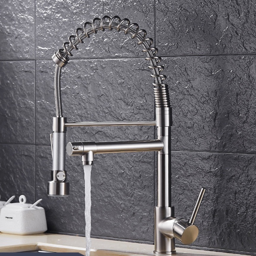 An allfixture Solid Brass Pull Out Spring Spout Kitchen Faucet with a flexible, coiled stainless steel spring neck and a black handle is installed against a dark tiled backsplash. The faucet features two spouts and a lever handle for temperature and flow control. A white jar sits on the counter in the background.