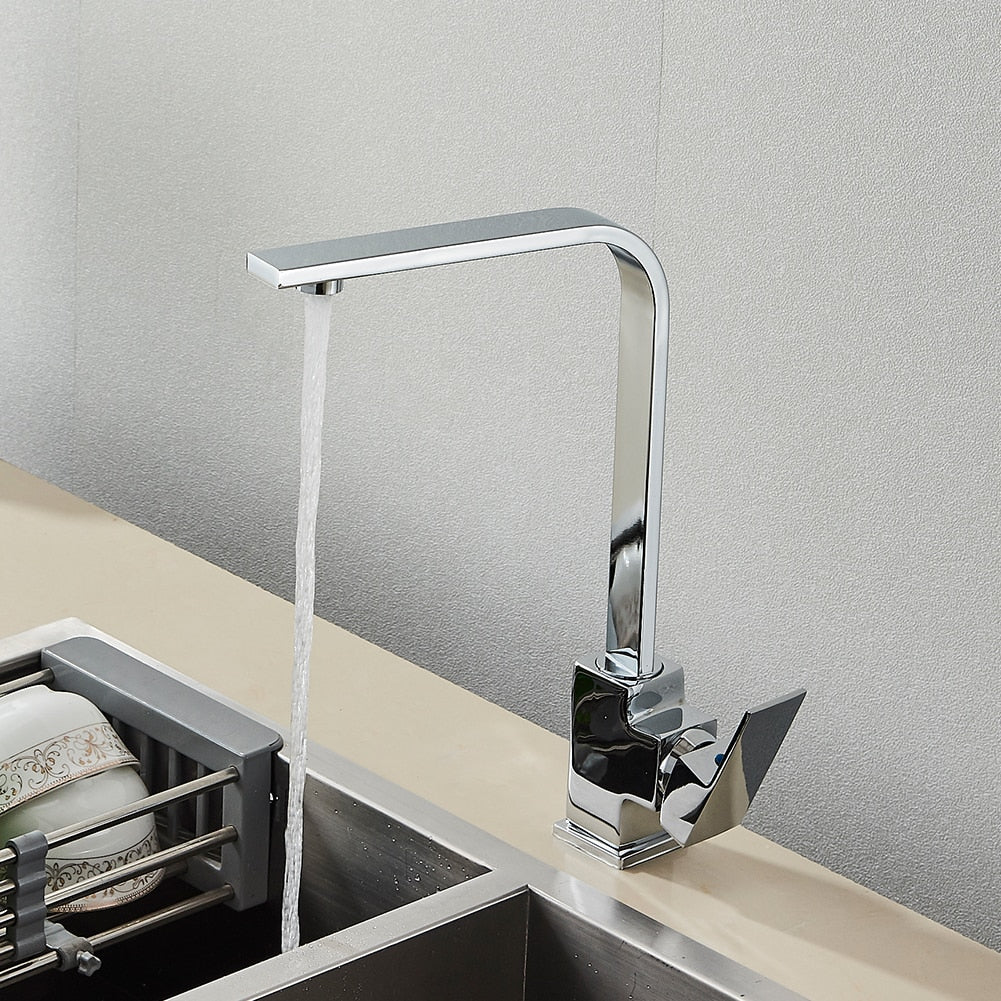 The Deck Mounted Single Handle Kitchen Faucet by Rozin, with its sleek and minimalist brushed gold finish, releases a steady stream of water into a stainless steel sink. It features a right-angled spout and an angular handle, set against a plain, light-colored wall.