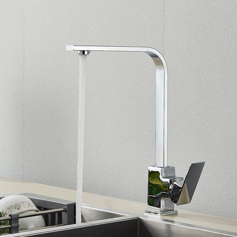 The Deck Mounted Single Handle Kitchen Faucet by Rozin, with its sleek and minimalist brushed gold finish, releases a steady stream of water into a stainless steel sink. It features a right-angled spout and an angular handle, set against a plain, light-colored wall.