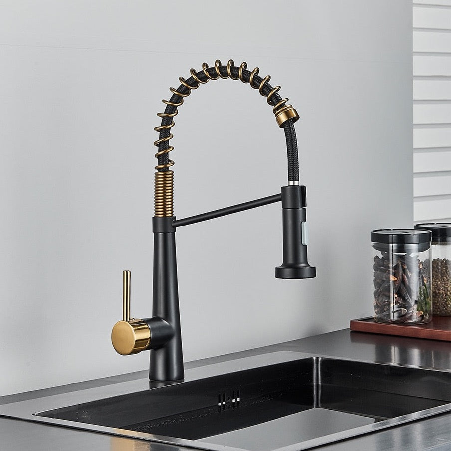 A modern Rozin Deck Mounted Pull Down Sprayer Kitchen Sink Faucet, featuring a sleek black finish with gold accents, is installed over a stainless steel sink. The faucet boasts a curved, flexible spring spout with a convenient pull-down sprayer. In the background, jars and a white panel on the wall enhance the sleek design.