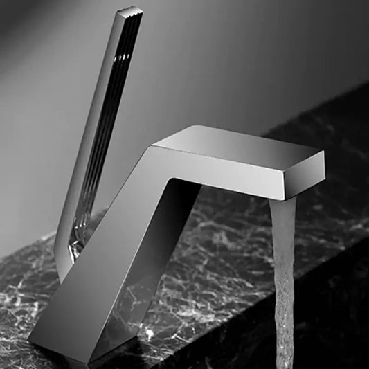 A modern, sleek Becola Single Handle Brass Bathroom Sink Faucet with an angular design pours water into a black sink. A blurred bottle and reed diffuser are in the background, set against a minimalist dark wall. The image is in black and white.
