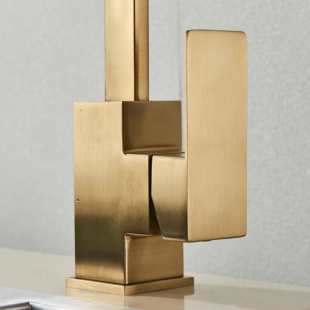 The Deck Mounted Single Handle Kitchen Faucet by Rozin, with its sleek and minimalist brushed gold finish, releases a steady stream of water into a stainless steel sink. It features a right-angled spout and an angular handle, set against a plain, light-colored wall.