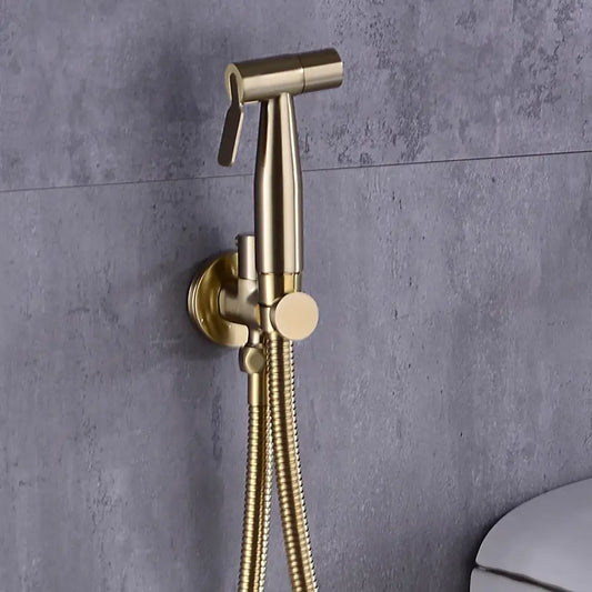 An allfixture Wasser™ Solid Brass handheld Toilet Sprayer Faucet, a modern wall-mounted, gold-colored bidet shower with a flexible hose, is installed against a gray textured wall. The handle is positioned upward, similar to a sleek bathroom sink faucet, and part of a white toilet is visible in the lower right corner.