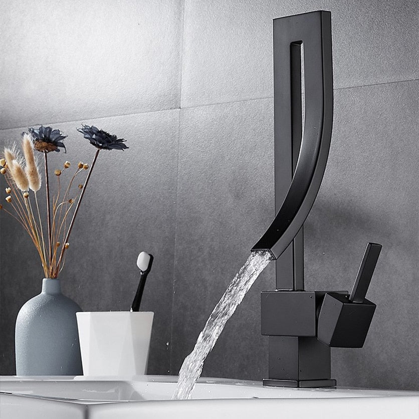 A modern, sleek Deck Mounted Brass Bathroom Basin Faucet from Bacala, featuring a curved design in black, is dispensing water into a white sink. In the background, there's a vase with dried flowers and a toothbrush holder containing a black and white toothbrush. The wall appears to be tiled in a dark color, echoing the elegance of a premium shower head.