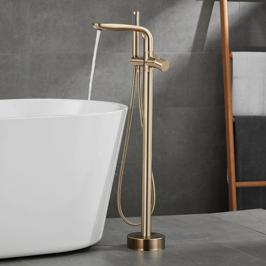 The Mike Jake Valmont Freestanding Bathtub Faucet With Hand Shower, featuring solid brass construction and a gold finish, stands by a white tub in a bathroom with gray tiled walls and a wooden towel rack.